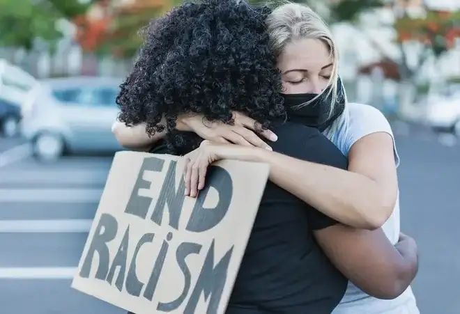 Two women hug, one has an "End Racism" banner in her hands. By Sabrina/stock.adobe.com