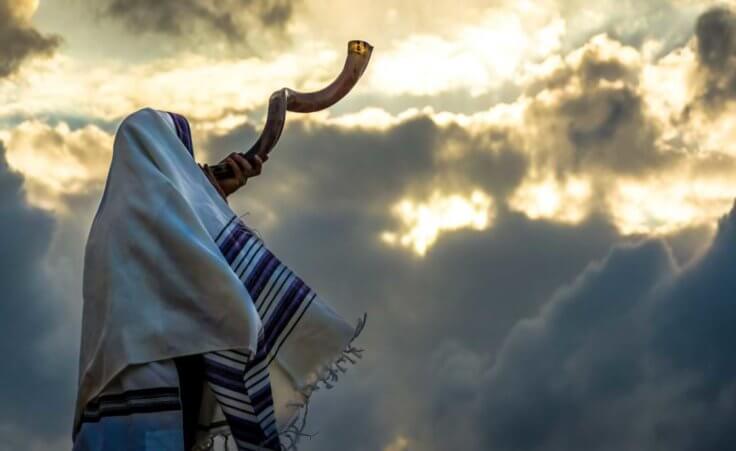 A Jewish man in a tallith prayer shawl blow's a shofar (ram's horn) against dramatic sky. © John Theodor/stock.adobe.com