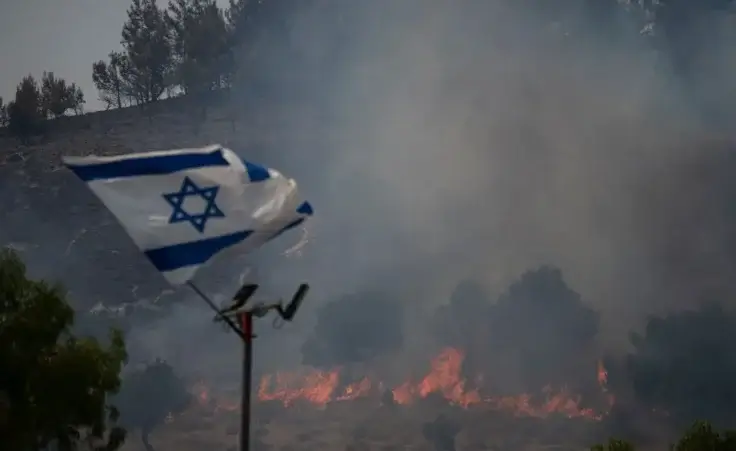 An Israeli flag flutters next to a fire burning in an area near the border with Lebanon, northern Israel in Safed, Wednesday, June 12, 2024. Scores of rockets were fired from Lebanon toward northern Israel on Wednesday morning, hours after Israeli airstrikes killed four officials from the militant Hezbollah group including a senior military commander. (AP Photo/Leo Correa)