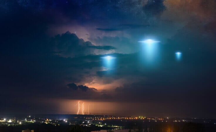 Extraterrestrial aliens spaceship fly above small town, UFO with blue spotlights in dark stormy sky. By IgorZh/stock/adobe.com