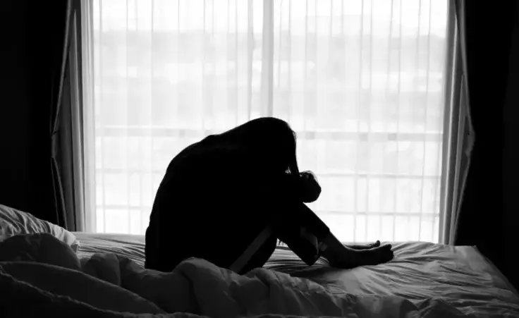 Silhouette of a woman sitting on the bed beside the windows with sunlight in the morning to illustrate feeling depressed. By Kenstocker/stock.adobe.com